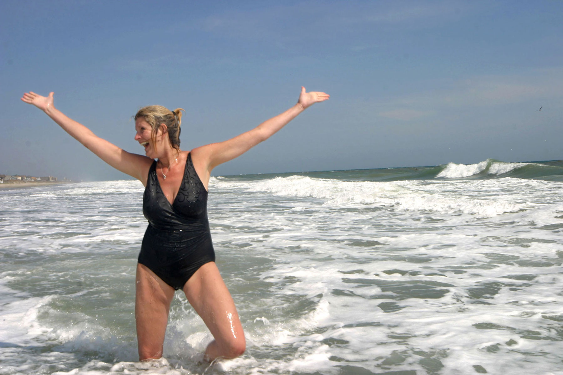 A woman in a black, one-piece bathing suit tosses her hands in the air while standing in shallow water