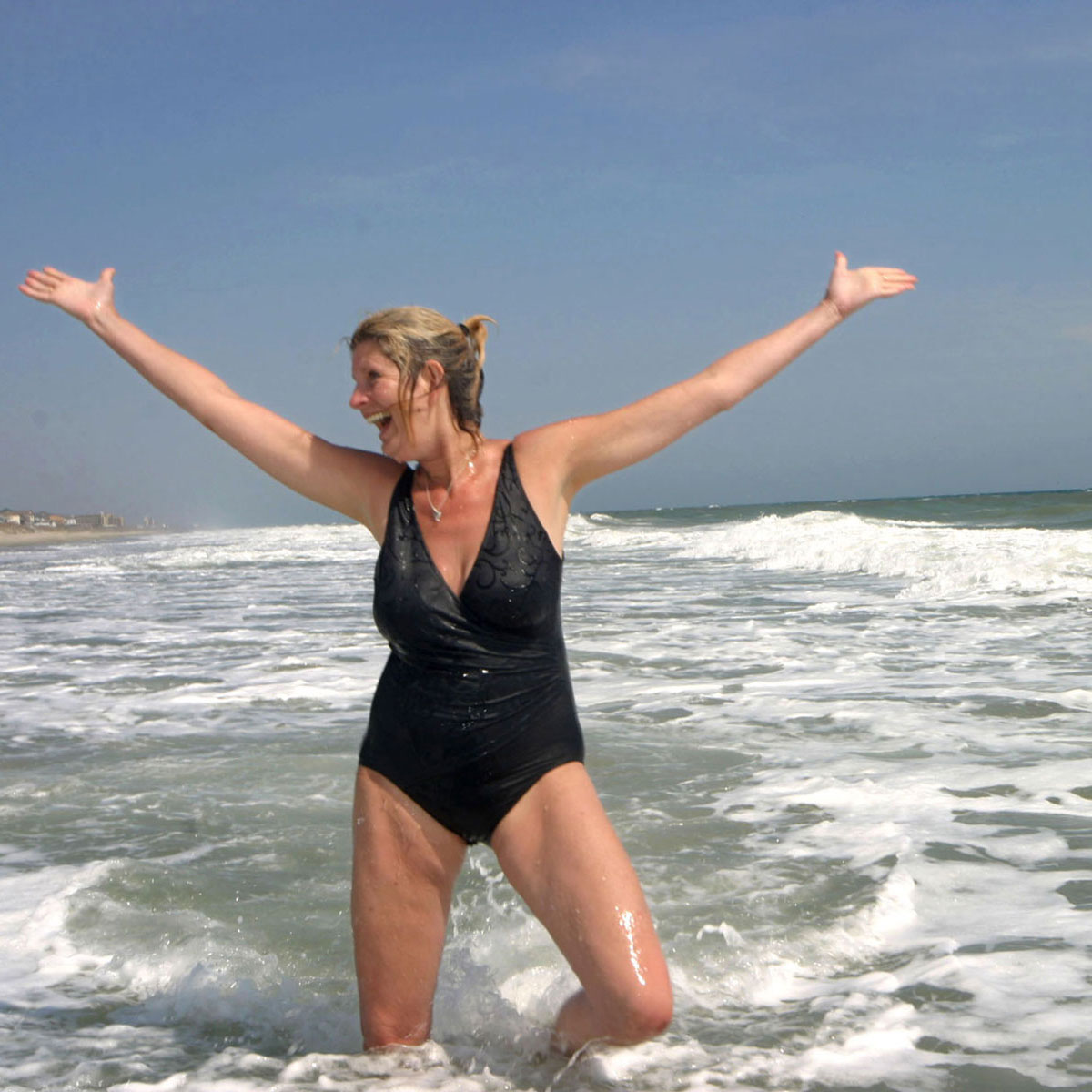 A woman in a black, one-piece bathing suit tosses her hands in the air while standing in shallow water