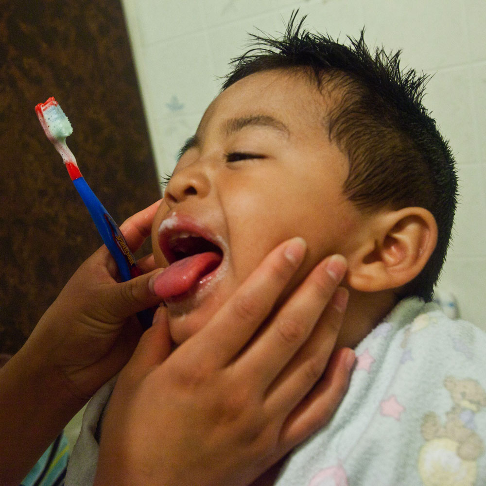 A boy sticks out his tongue while his mom brushes his teeth