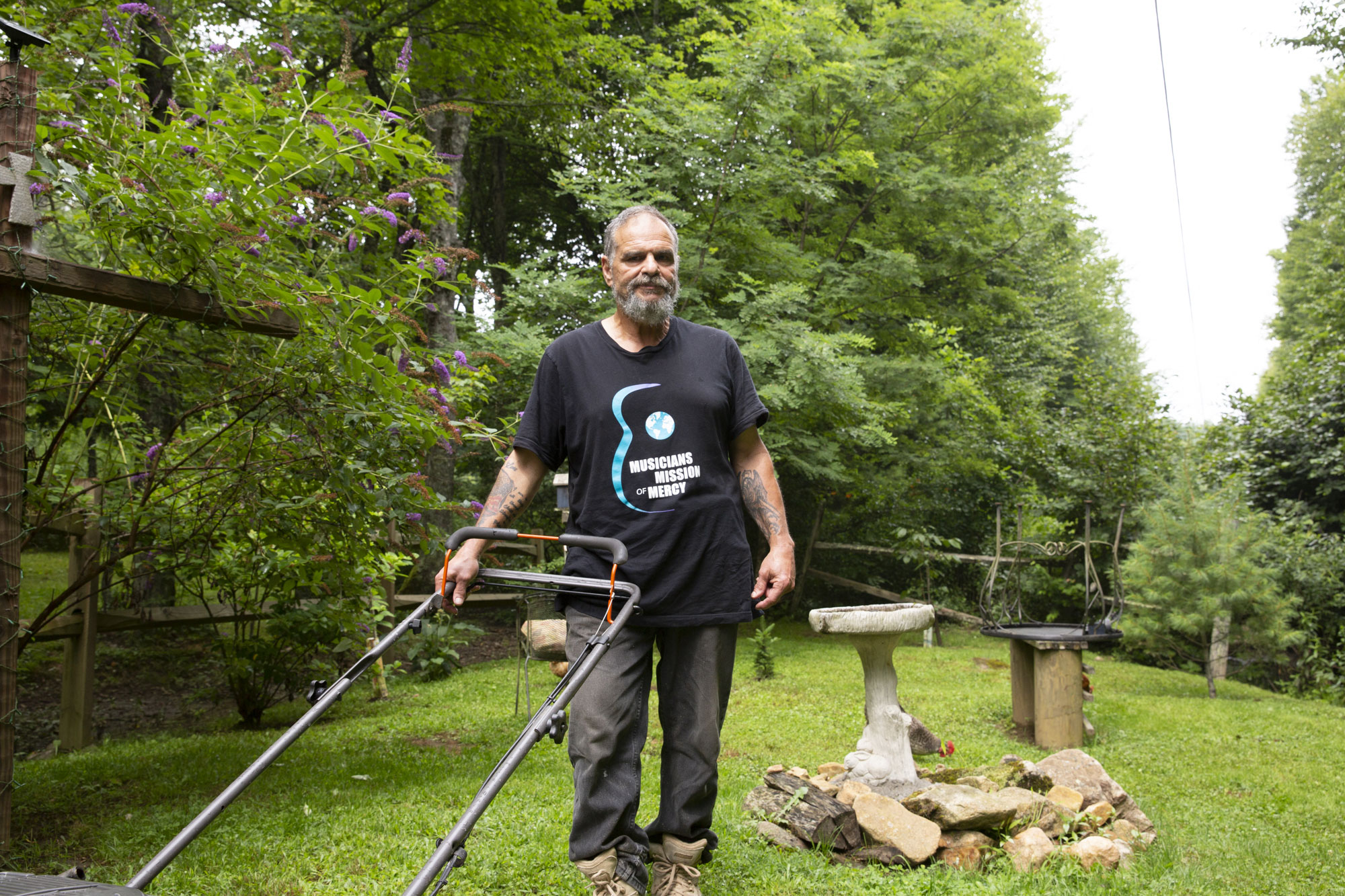Gray haired man stands next to his lawnmower in a garden
