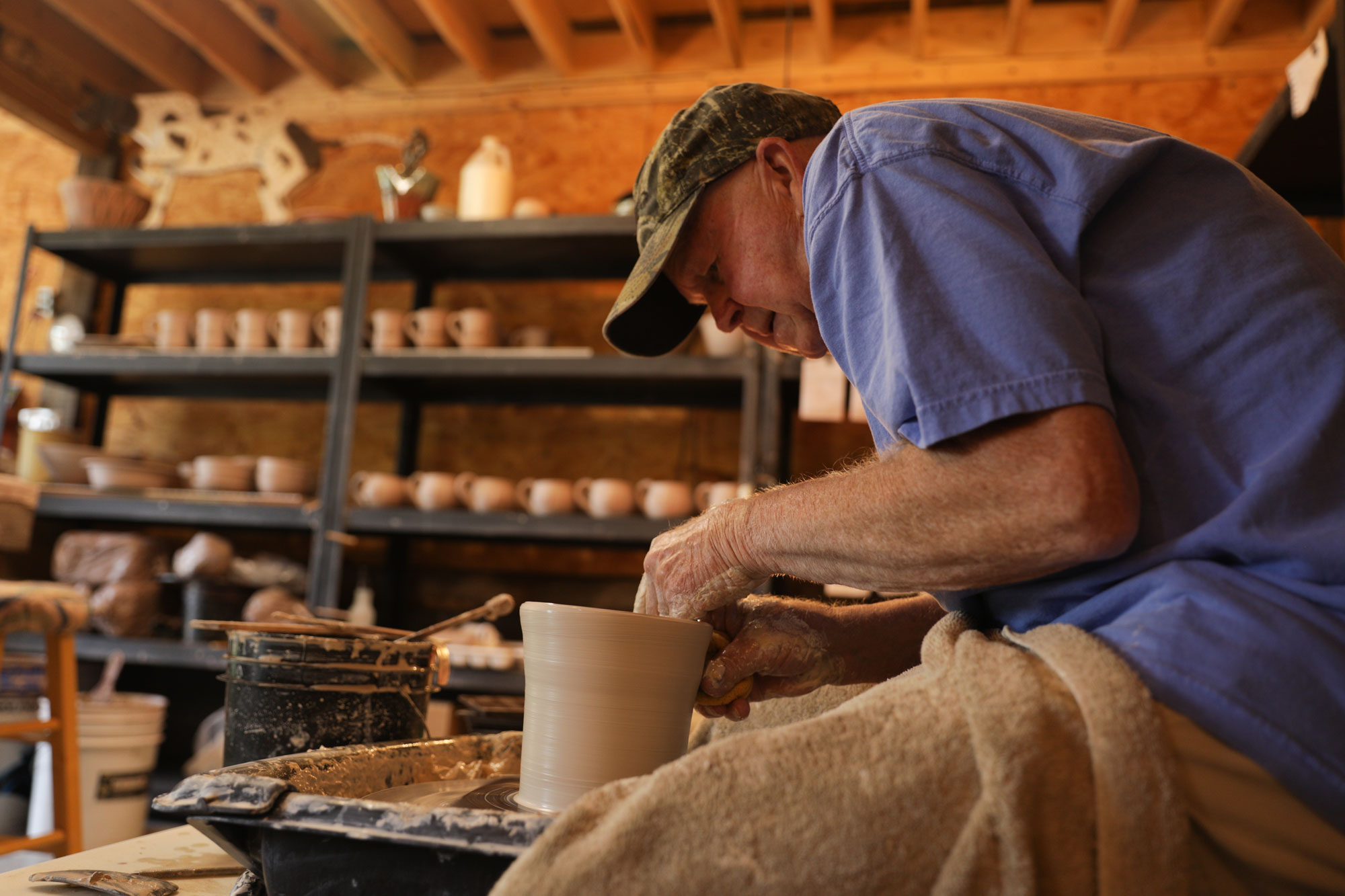 An older man leans over a potters wheel as he makes a utensil holder