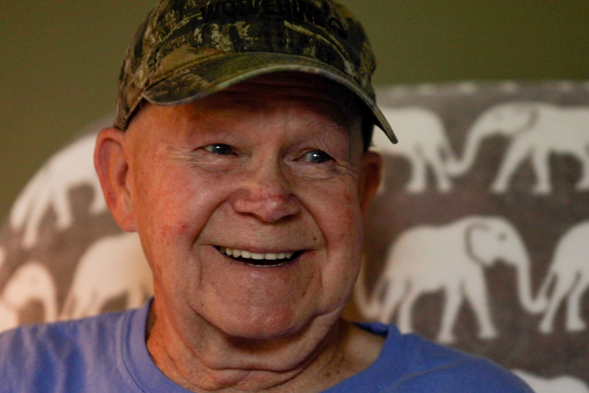 An older man with a camo hat on smiles at the camera