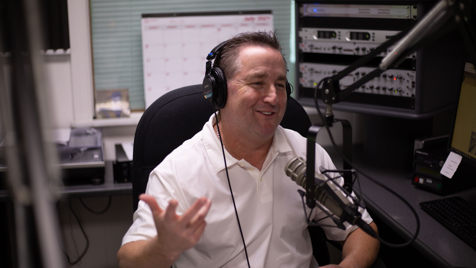 A man in a white polo shirt wears headphones and speaks into a microphone