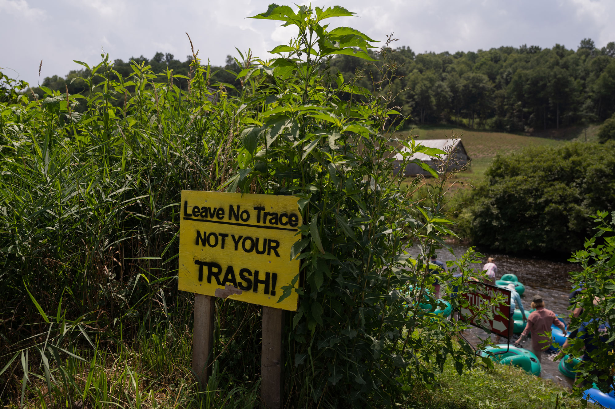 A sign saying "Leave no trace, not your trash!" sits in front of a river where people are tubing