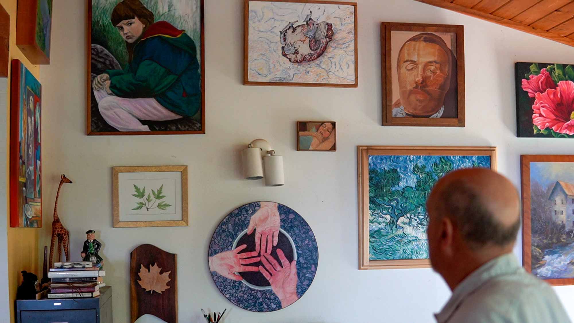 A man looks at his framed paintings covering his wall