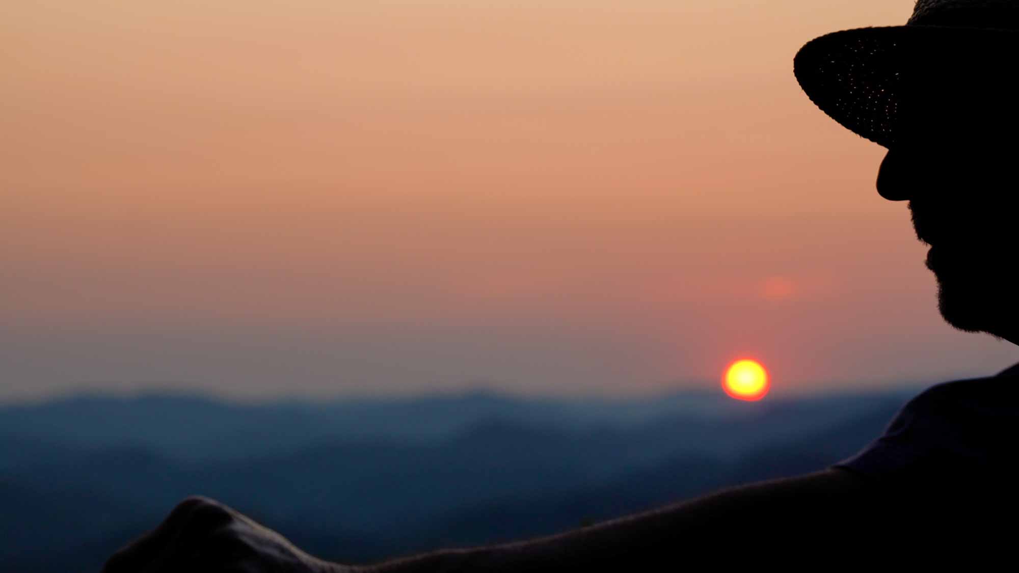A man's silhouette sits before a sun setting over distant mountains