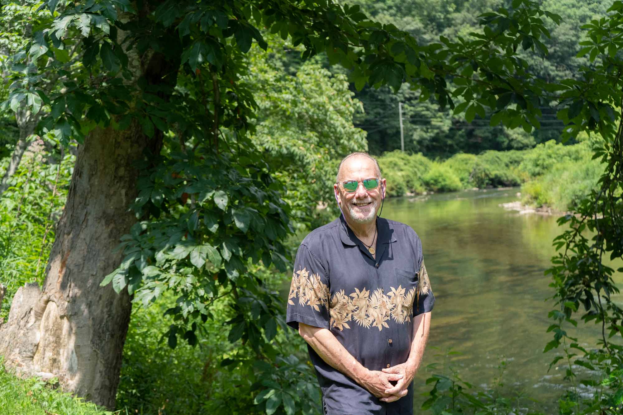 Man in button down black shirt and sunglasses stands by a river