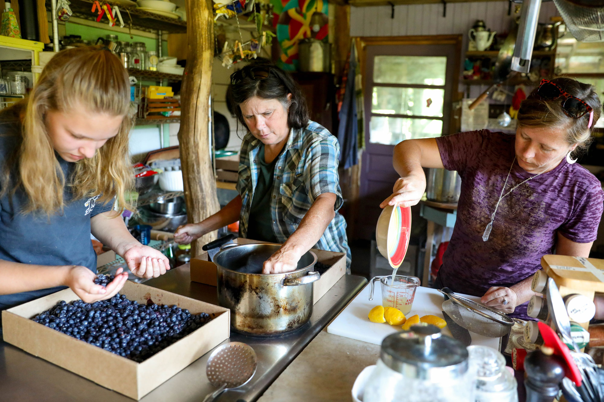 Three women prepare blueberry syrup, gathering berries, putting them in a pot, and pouring lemonade