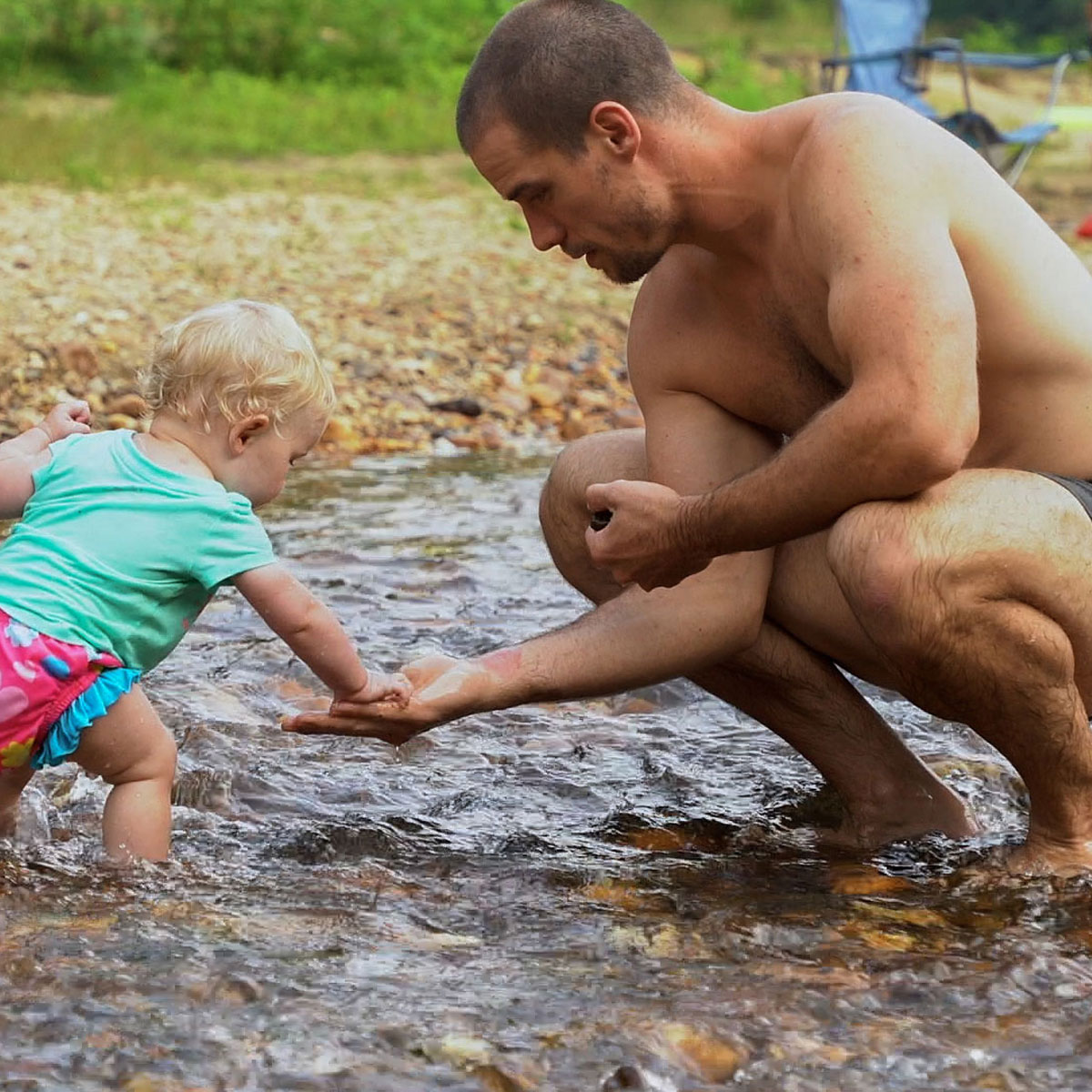 A shirtless man holds out a stone to a little girl in the water