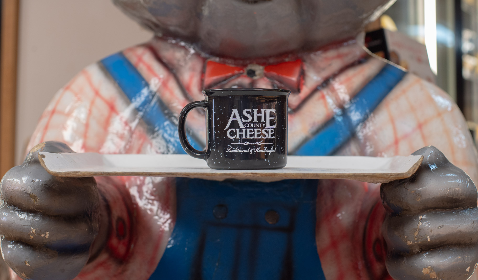 A mug saying "Ashe County Cheese" sits on a display tray held up by a ceramic animal