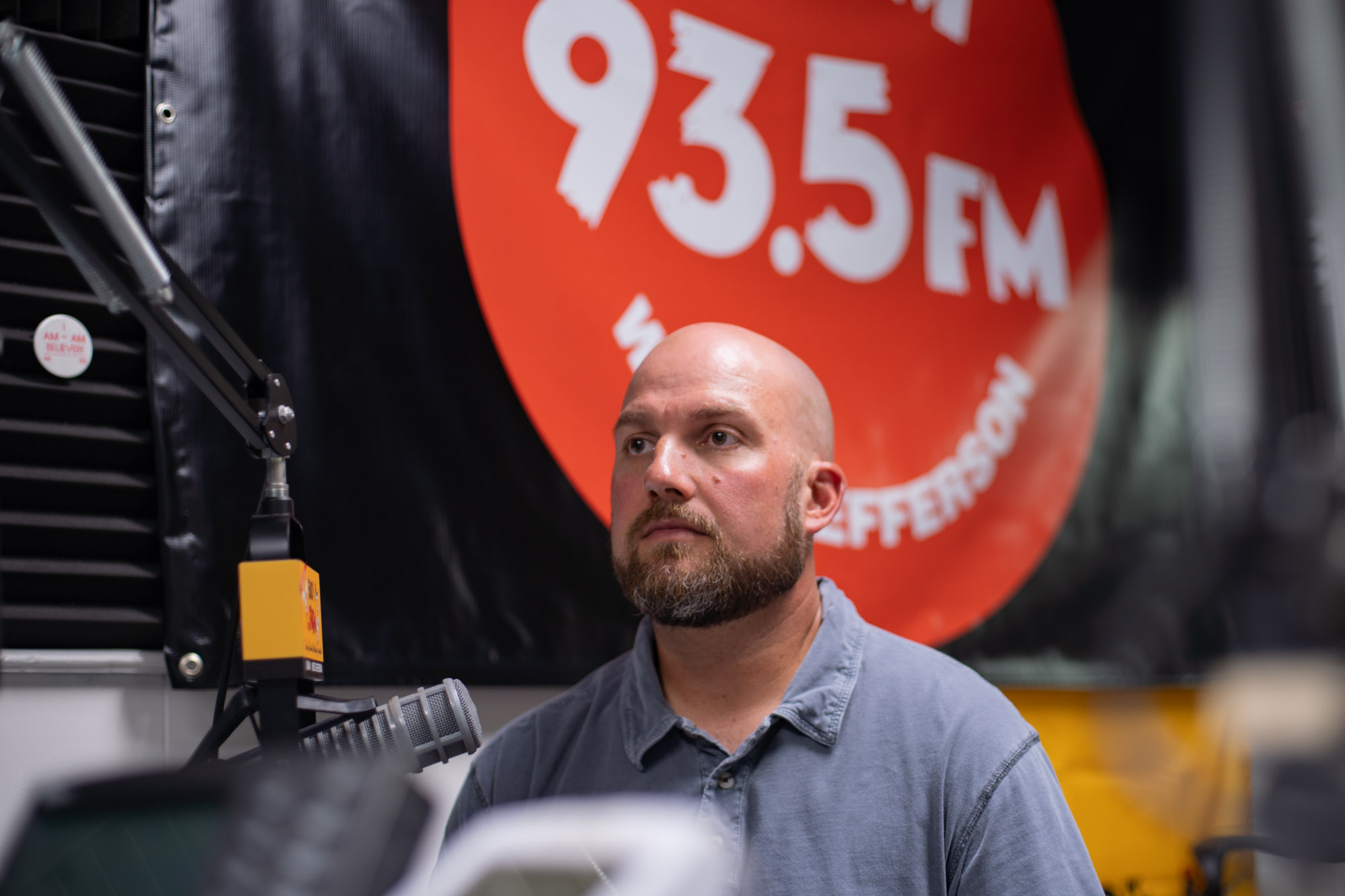 A bald man with a beard sits in front of radio station microphone